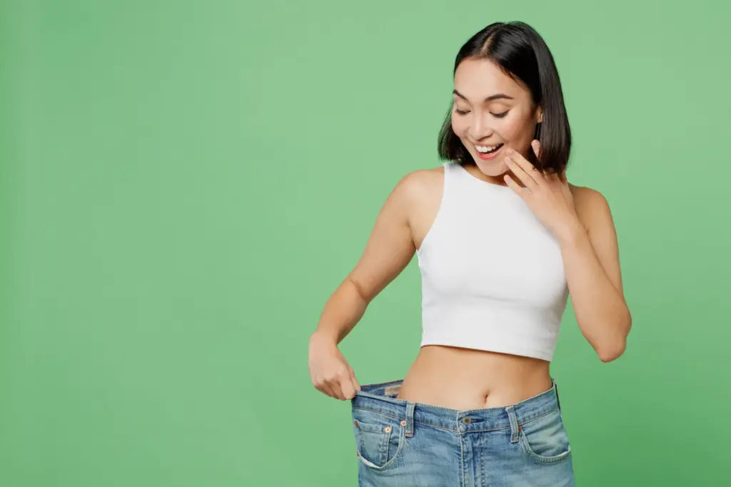 Young happy woman wears white clothes and show loose pants on waist after weight loss in Salt Lake City, UT | Santosh Yoga Institute